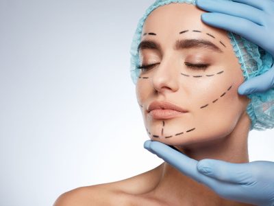 Beauty portrait of woman with closed eyes, plastic surgery concept, studio. Head and shoulders of model in blue cap with puncture lines on face, hands in blue gloves holding face